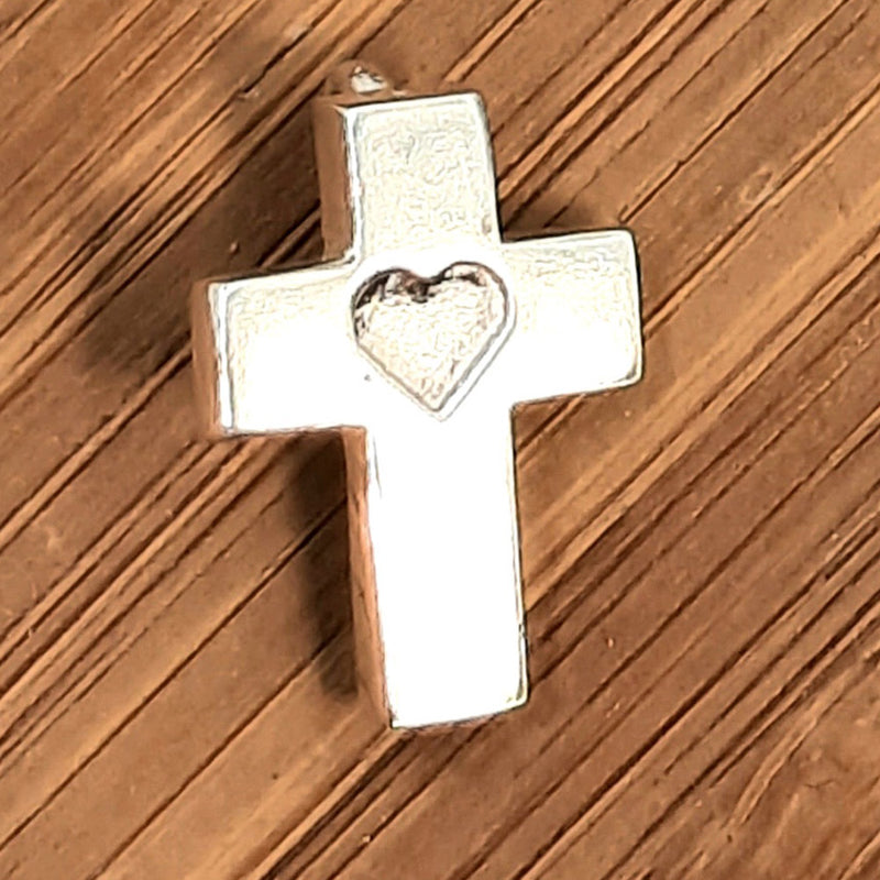 small cross around 20mm high and 14mm across. At the top there is a small u ring to which a jump ring is attached. A shape of a heart is engraved in the intersection between the vertical and horizontal posts. The outline of the cross is approsimately 3mm wide and 1mm thick. The finish is shiny. The cross is shown on a piece of wood.